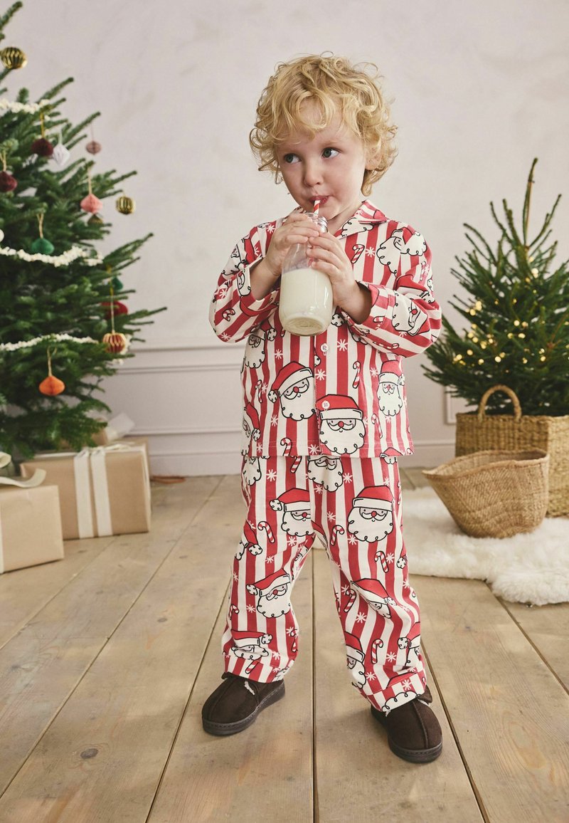 Rot-weiße gestreifte Schlafanzüge mit Mustern von Weihnachtsmann und Zuckerstangen, aus weichem Stoff gefertigt. Ein Kind hält eine Flasche Milch.
