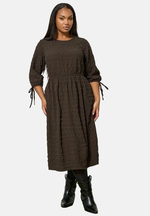 Vrouw in een donkergroene textuur midi-dress met 3/4 mouwen die bij de manchetten zijn gestrikt, gecombineerd met zwarte panty's en zwarte cowboylaarzen.