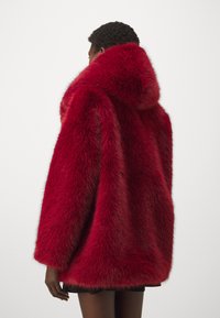 Rød faux fur-kåpe med hette, med en fluffy tekstur og en løs passform. Ermene er brede og nederkanten er litt avrundet.