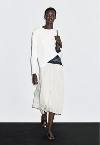 Hvid langærmet skjorte og en flydende, cremet nederdel, kombineret med et marineblåt tørklæde og brune lædersandaler. Lille sort taske i hånden.