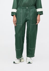 Osoba stojaca v rozvolnených tmavozelených nohaviciach s kontrastným šitím a bielych teniskách, uvoľnene postavená na jednoduchom pozadí.