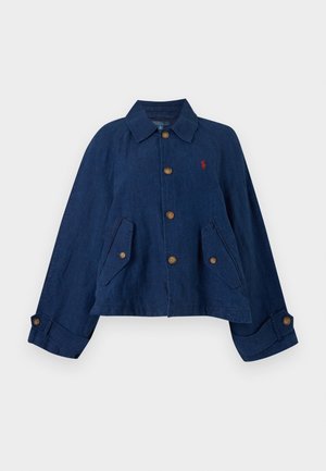 Kvinde står iført en mørkeblå jakke over en lyseblå skjorte, falmede blå jeans, hvide sneakers og holder en lille brun håndtaske.