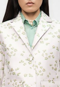 Κοντινή λήψη ατόμου που φοράει λευκό σακάκι με πράσινο λουλουδένιο σχέδιο, πράσινο πουκάμισο με κουμπιά και πολύστιχη μαργαριταρένια κολιέ.