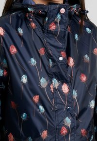 Giacca impermeabile blu navy con motivo floreale con fiori rossi, rosa e blu, cappuccio e bottoni a contrasto. Texture liscia e lucida.