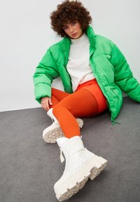 Chaqueta acolchada verde, suéter blanco, leggings naranjas y botas blancas gruesas. Materiales texturizados con un acabado brillante y detalles estampados.