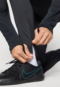 Sorte atletiske leggings med lynlås ved anklen og tætsiddende design, kombineret med sorte sko med et grønt Nike-logo og hvide sokker.