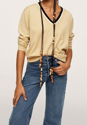 Personne portant un pull beige à col en V, un jean bleu taille haute et de longues tresses décorées de perles en bois tombant devant.