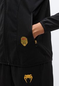 Μαύρο αθλητικό μπουφάν με υφή, με χρυσλά λογότυπα UFC και Venum, τσέπες με φερμουάρ και ελαστικά μανίκια.