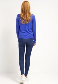 Modrý svetr s dlouhými rukávy, přiléhavý střih, doplněný tmavými úzkými džínami a bílými teniskami. Vlasy padají vlnitě přes ramena.