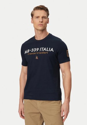 Uomo che indossa una t-shirt MB-339 Italia blu navy con scritta ed emblema arancione, abbinata a pantaloni beige, in piedi davanti a uno sfondo semplice.