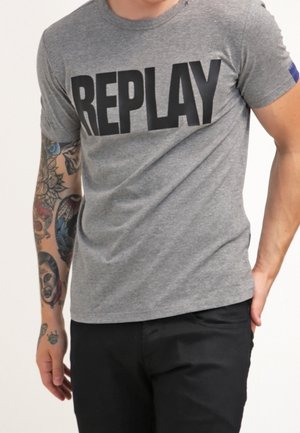 Grå bomuld T-shirt med et stort sort "REPLAY" print på fronten. Korte ærmer og rund halsudskæring. Sorte bukser bæres nedenunder.