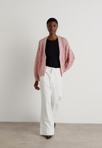 Cardigan rosa lavorato a maglia con una vestibilità ampia e un motivo testurizzato, indossato sopra a una maglietta nera e abbinato a pantaloni bianchi a gamba larga.