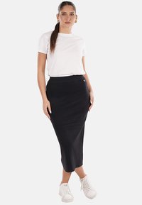 Una donna in piedi con i capelli lunghi raccolti, che indossa una t-shirt bianca, una gonna midi nera aderente e sneakers bianche su uno sfondo semplice.