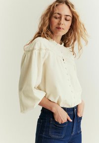 Bluzka w kolorze off-white z teksturowym wykończeniem, z falbaniastym dekoltem, zapinana na guziki, z bufiastymi rękawami, w zestawieniu z dżinsami z wysokim stanem.