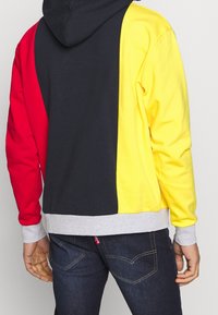 Mikina s kapucí s barevným blokovým designem, obsahující červené, černé a žluté panely. Šedé manžety a lem, vyrobená z měkkého materiálu.
