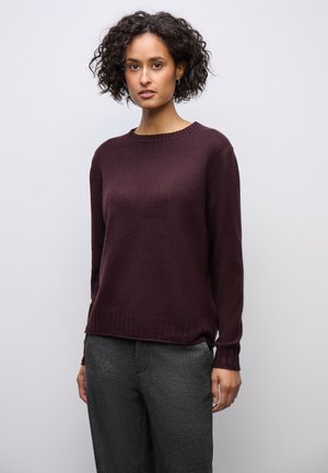 Vrouw met krullend haar, gekleed in een donkerpaarse gebreide trui en grijze broek, staand tegen een effen lichte achtergrond.