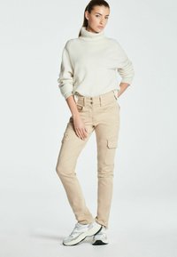 Beige cargo kalhoty s bočními kapsami, doplněné bílým rolákem. Model má na sobě šedé a bílé tenisky. Jednoduché pozadí.