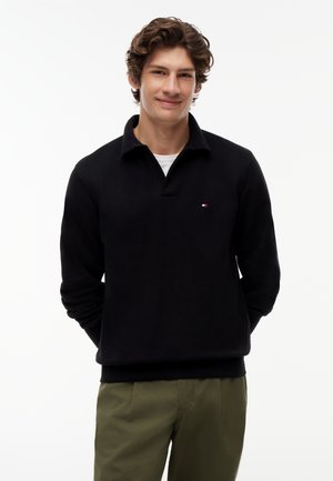 Ung mand smilende, iført en sort langærmet pullover med et lille logo og olivengrønne bukser, stående med hænderne bag ryggen.