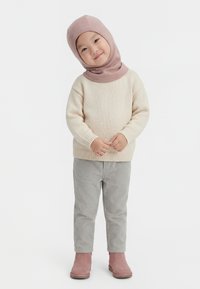 Kind draagt een beige trui, grijze broek, roze laarzen en een lichtroze capuchon, staat met gevouwen handen en glimlacht licht.