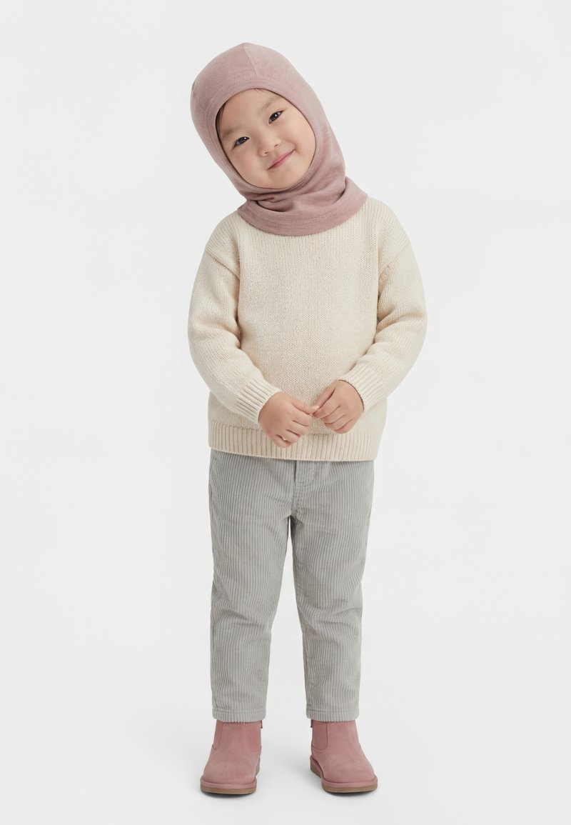 Kind draagt een beige trui, grijze broek, roze laarzen en een lichtroze capuchon, staat met gevouwen handen en glimlacht licht.