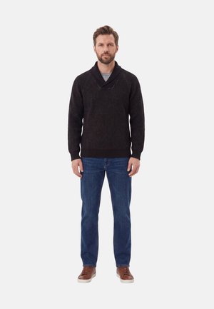 Uomo in piedi di fronte, indossa un maglione scuro, jeans blu e scarpe marroni su uno sfondo chiaro uniforme.