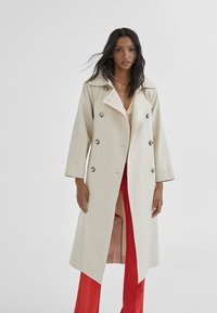 Beżowy płaszcz trench z podwójnym zapięciem, dużymi klapami i dwiema bocznymi kieszeniami. Posiada ciemne akcenty guzikowe i luźny krój.