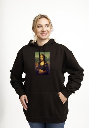 Smilende kvinde med blondt hår iført sort hættetrøje med Mona Lisa-tryk, hænder i lommerne, stående mod hvid baggrund.