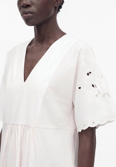 Personne à la peau foncée portant une robe blanche à col en V avec de larges manches présentant des détails découpés floraux brodés.