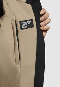 Beżowa i czarna podszewka kurtki z naszywką z napisem "Khujo" w białym kolorze. Gładki materiał z kieszenią po lewej stronie.