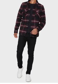 Musta ja burgundipunainen ruudullinen takki, jossa on kaksi rintataskua, valmistettu kankaasta, ja jossa on napitettavat etuosat sekä pitkät hihat. Päällä musta paita.