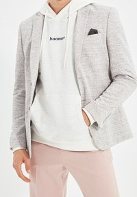 Blazer grigio chiaro con un motivo testurizzato su una felpa bianca con il testo "boomer". Abbinato a pantaloni rosa chiaro e un fazzoletto da taschino nero.
