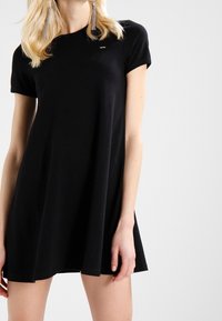 Černé krátké šaty s volným střihem, vyrobené z měkké látky. Mají malé logo na hrudi a sahají těsně nad kolena.
