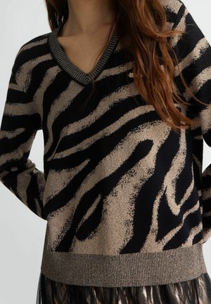 Femme portant un pull à col en V rayé zèbre noir et or, manches longues et ourlet côtelé métallique.