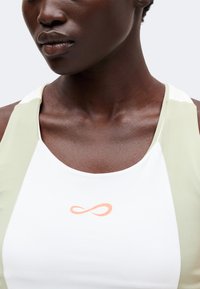 Gros plan d'une personne portant un haut de sport blanc et vert clair avec un petit symbole d'infini orange sur la poitrine.