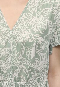 Detailný záber osoby oblečenej v zelenej a bielej šaty s kvetinovým vzorom, výstrihom do V, krátkymi rukávmi a zoševnou páskou.