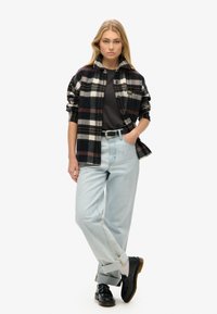 Camicia flanella a quadri bianco e nero sopra una maglietta scura, abbinata a jeans azzurri chiari e scarpe nere. Modello in piedi con le mani in tasca.