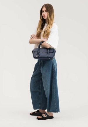 Jeune femme aux longs cheveux debout, portant une chemise blanche, un jean bleu à jambes larges, des sandales noires, tenant un sac à main Picard bleu marine.