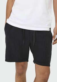 Pantalones cortos negros de tejido con cinturilla elástica y cordón. Cuenta con bolsillos laterales y una tela texturizada. Combinado con una camiseta blanca.