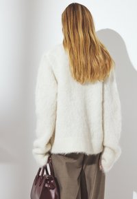 Vit fluffig tröja med oversizeärmar, kombinerad med beiga byxor och en liten mörk vinröd handväska. Mjuk textur och avslappnad passform.