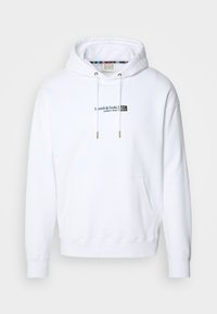 Felpa bianca con tasca a marsupio, cappuccio con coulisse e dettagli dorati. Presenta il logo "Scotch & Soda" in blu navy e un tessuto texturizzato.