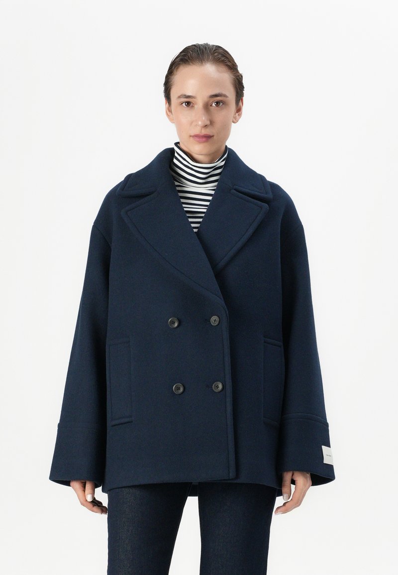 Pardessus peacoat in lana blu navy con design doppiopetto, ampie revers, bottoni e tasche laterali; indossato sopra un dolcevita a righe bianche e nere.