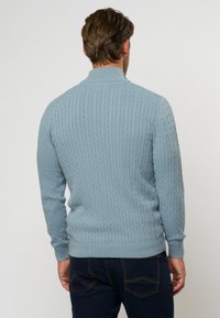 Šviesiai mėlynas nertinis megztinis su apykakle ir raštuotomis rankogaliais bei apačia. Tekstūra yra storas, su pakeltu raštu visame megztinyje.