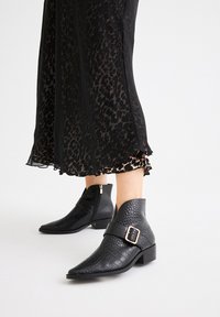 Botas negras hasta el tobillo con un acabado texturizado similar al cocodrilo. Punta afilada y cremallera lateral. Una correa con hebilla añade detalle.