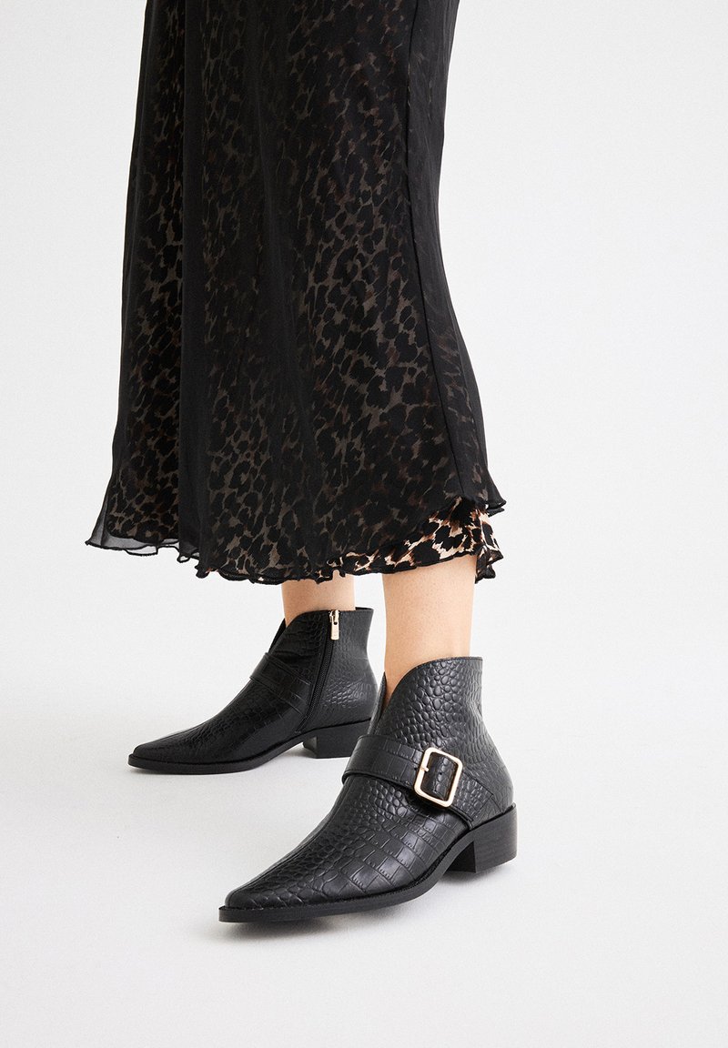 Botas negras hasta el tobillo con un acabado texturizado similar al cocodrilo. Punta afilada y cremallera lateral. Una correa con hebilla añade detalle.