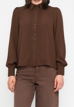 Femme portant une chemise marron à manches longues boutonnée avec un détail plissé à l'avant et un pantalon marron assorti, debout avec les bras détendus le long du corps.