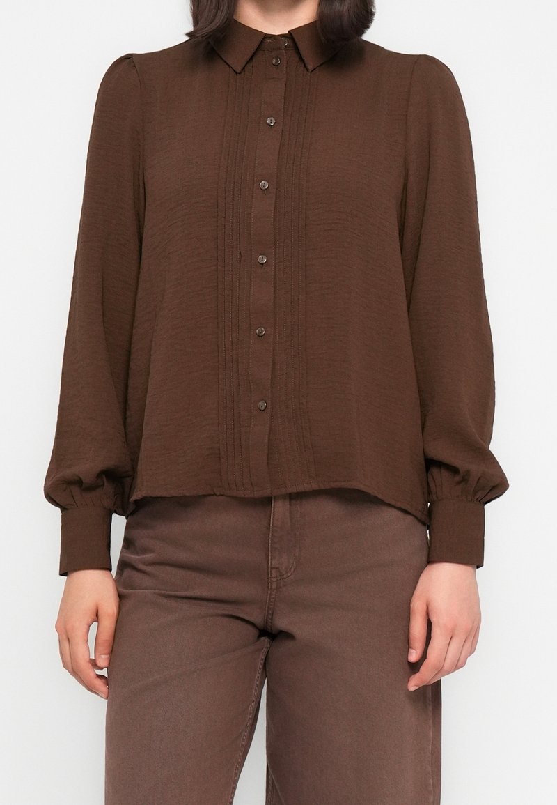 Femme portant une chemise marron à manches longues boutonnée avec un détail plissé à l'avant et un pantalon marron assorti, debout avec les bras détendus le long du corps.