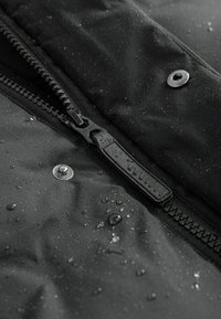 Gros plan d'une veste noire imperméable mettant en avant une fermeture éclair, un bouton à pression et des gouttes d'eau sur une surface lisse et texturée.