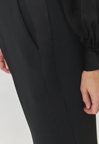 Abito in tessuto nero con una texture liscia, caratterizzato da una leggera lucentezza. Include una mano poggiata sul fianco, evidenziando il design.