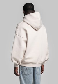 Cremefarvet hoodie med en stor, rund hætte, sænkede skuldre og ribbede manchetter. Blød tekstur, løs pasform og synlige sømdetaljer.