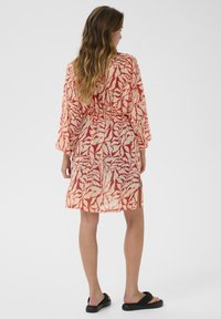 Vestito kimono trasparente con un motivo astratto rosso e crema, vita arricciata e maniche ampie, indossato con sandali neri.